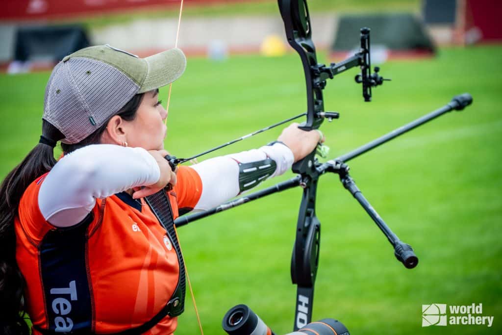 Een vrouw in een oranje shirt en een groene pet richt een samengestelde boog tijdens een handboogschietevenement buiten. Het World Archery-logo is zichtbaar in de hoek van de afbeelding.