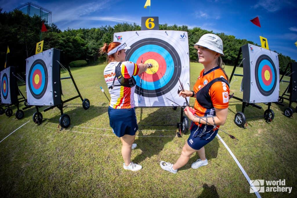 Twee boogschutters staan bij een doelwit met pijlen in de roos. Eén boogschutter wijst naar het doel terwijl de ander observeert. Doelen in baan 5 en 7 zijn ernaast zichtbaar. Het World Archery-logo staat in de hoek en benadrukt hun uitzonderlijke vaardigheden op het gebied van handboogschieten.