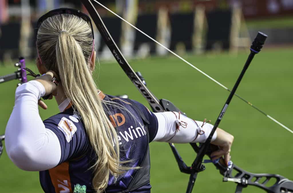 Een vrouwelijke boogschutter met lang blond haar, die sportkleding en een zonnebril draagt, bereidt zich voor om een pijl af te schieten vanuit haar compoundboog tijdens een handboogsportevenement in de open lucht.