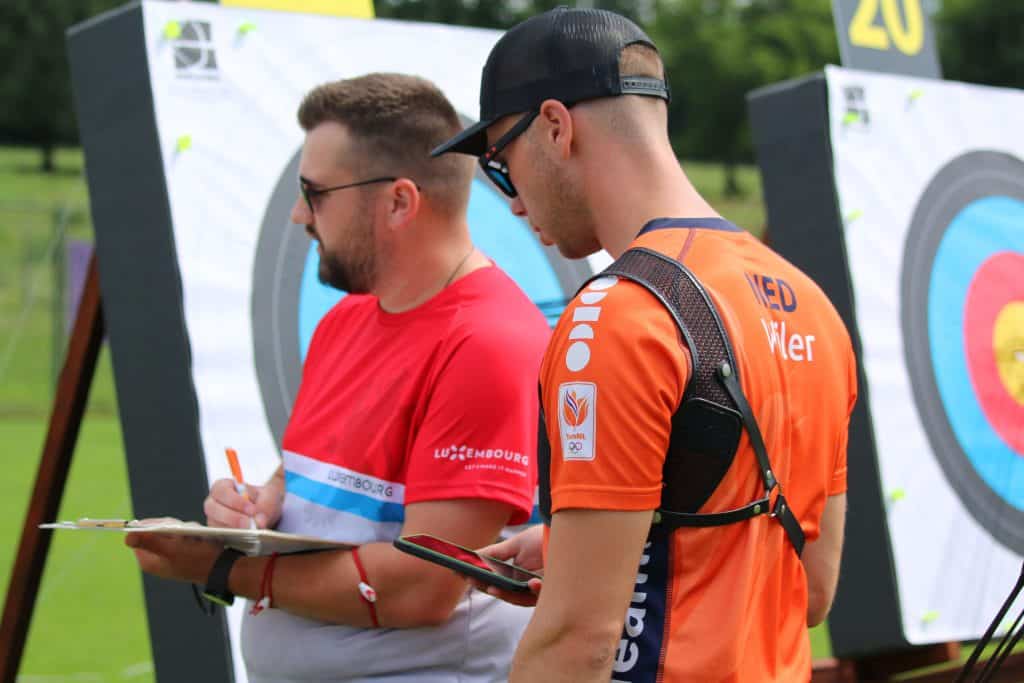 Twee boogschutters in wedstrijdkleding reviewscores naast boogschietdoelen met gekleurde ringen op een grasveld. De een draagt een rood shirt, de ander een oranje shirt met het opschrift "NED", beiden gericht op het perfectioneren van hun handboogschiettechniek.