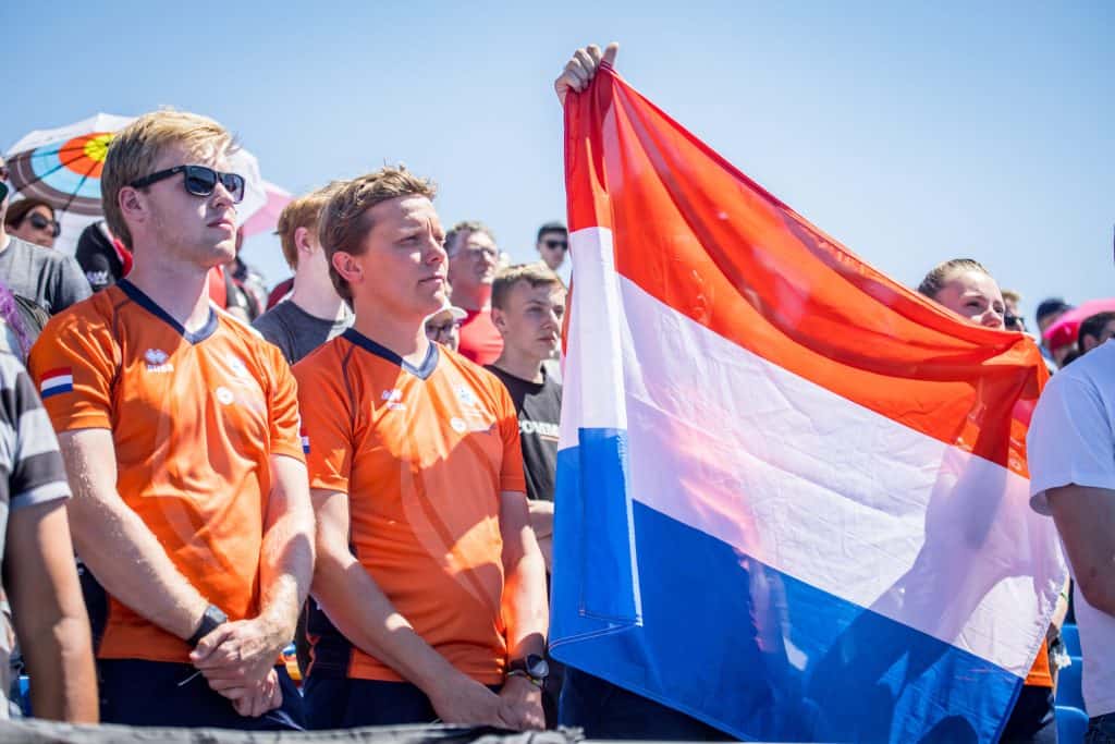 Mensen die oranje shirts dragen, houden een driekleurige vlag met rode, witte en blauwe horizontale strepen vast tijdens een handboogsportevenement in de open lucht.