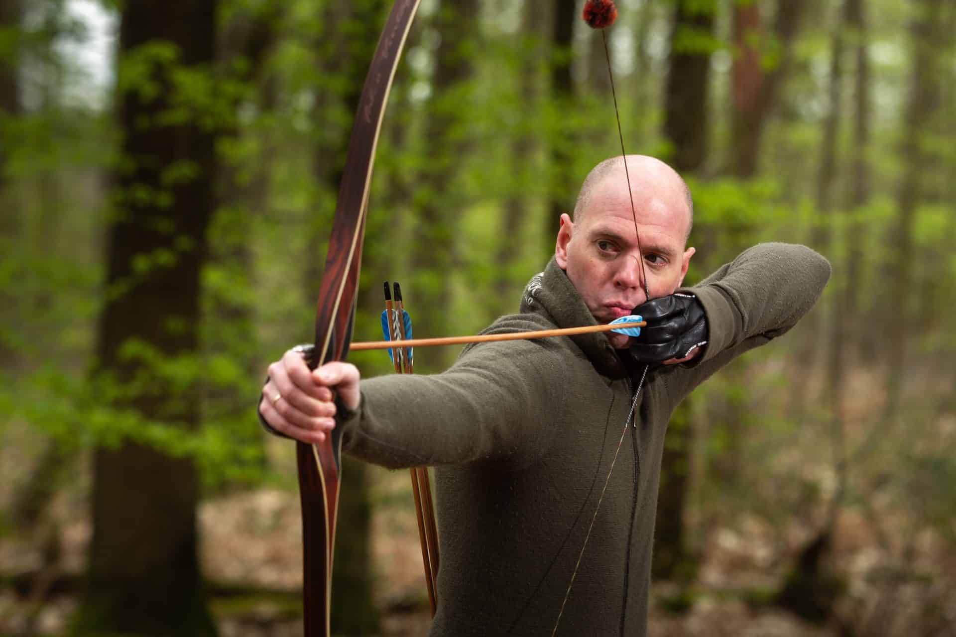Een persoon met een kaal hoofd richt een pijl en boog in een bosrijke omgeving, geconcentreerd op het doel, wat de precisie van handboogsport belichaamt.