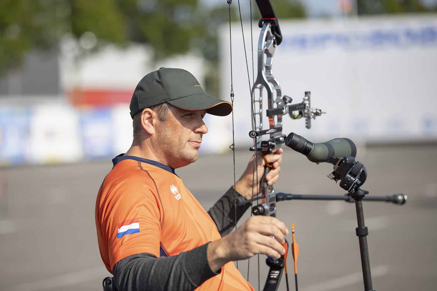 Een man met hoed, oranje shirt met een klein vlaggetje op de mouw, houdt een compoundboog vast en bereidt zich voor op handboogschieten. Hij is buiten met een onscherpe achtergrond van bomen en een gebouw.