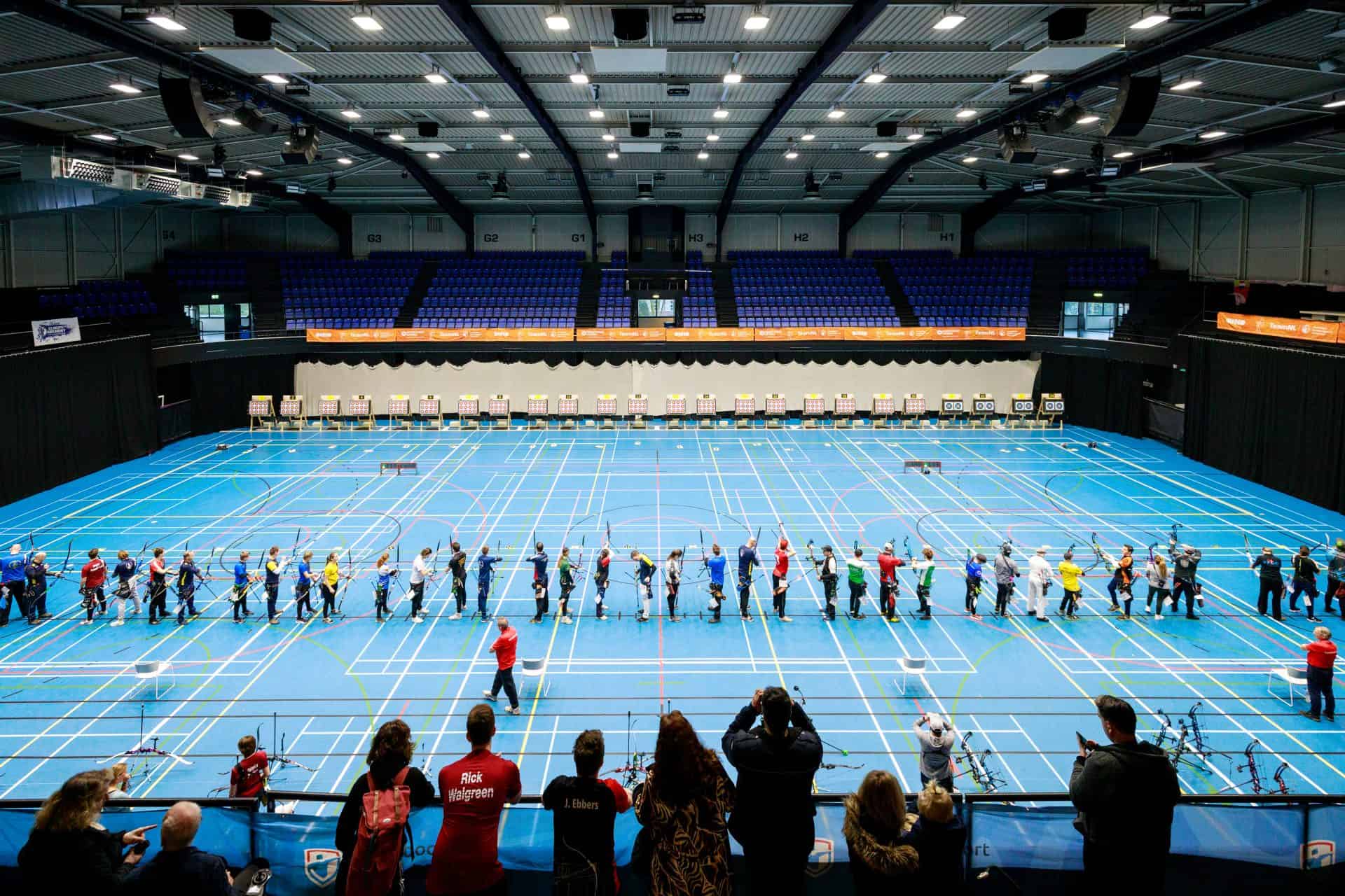 Een groep boogschutters staat in een rij en richt hun boog op doelen in een overdekte sporthal. Op de voorgrond zijn toeschouwers zichtbaar die gretig naar het handboogsportevenement kijken.