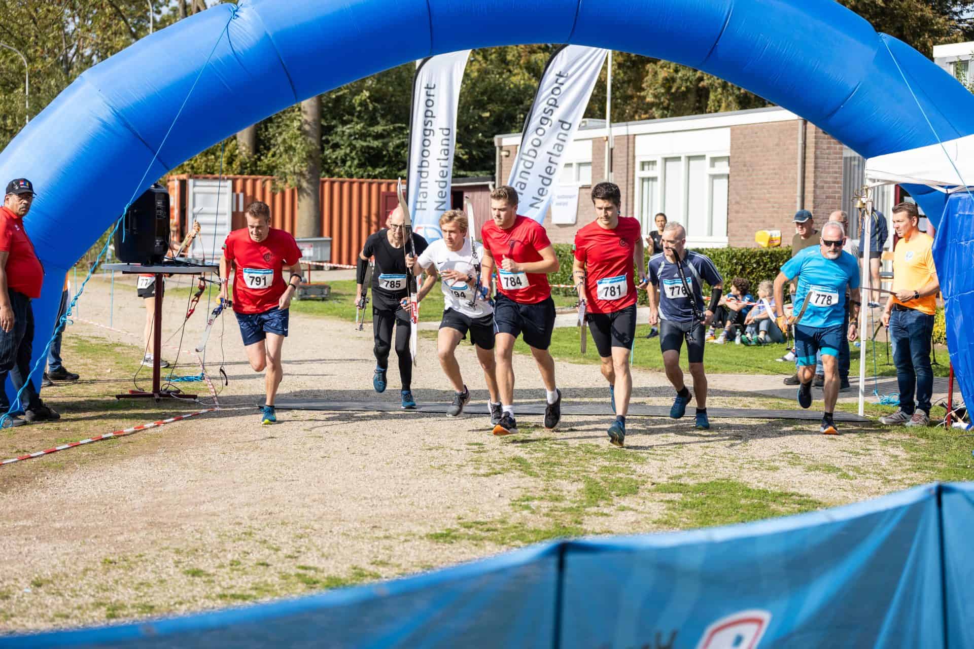 Een groep lopers begint een race onder een blauwe opblaasbare boog, met op de achtergrond enkele toeschouwers en een persoon in een geel shirt met een pijl.