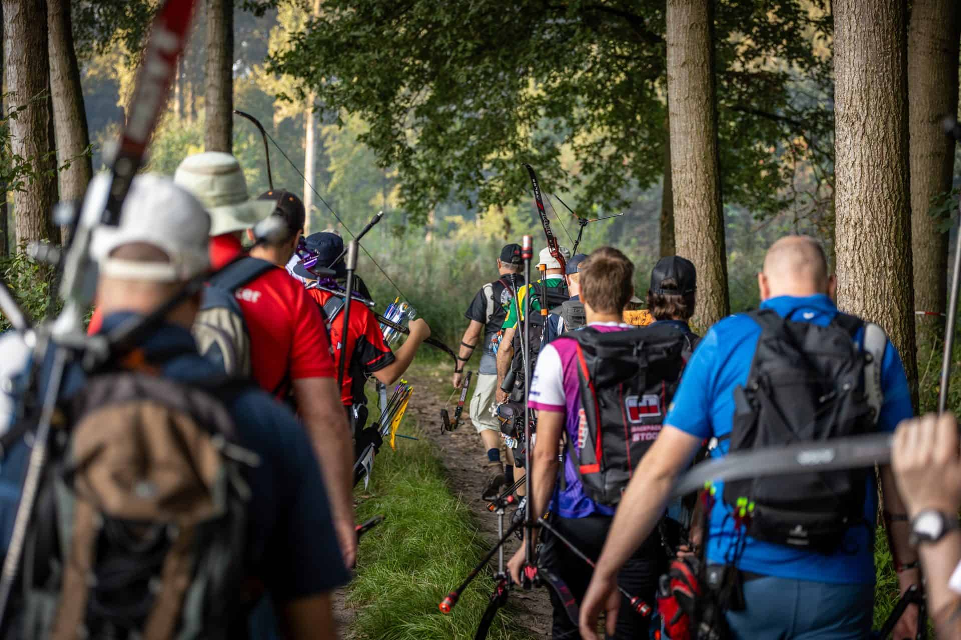 Een groep boogschutters loopt met bogen en uitrusting over een bospad, op weg naar hun bestemming voor een handboogsport. Bomen en groen omringen het pad.