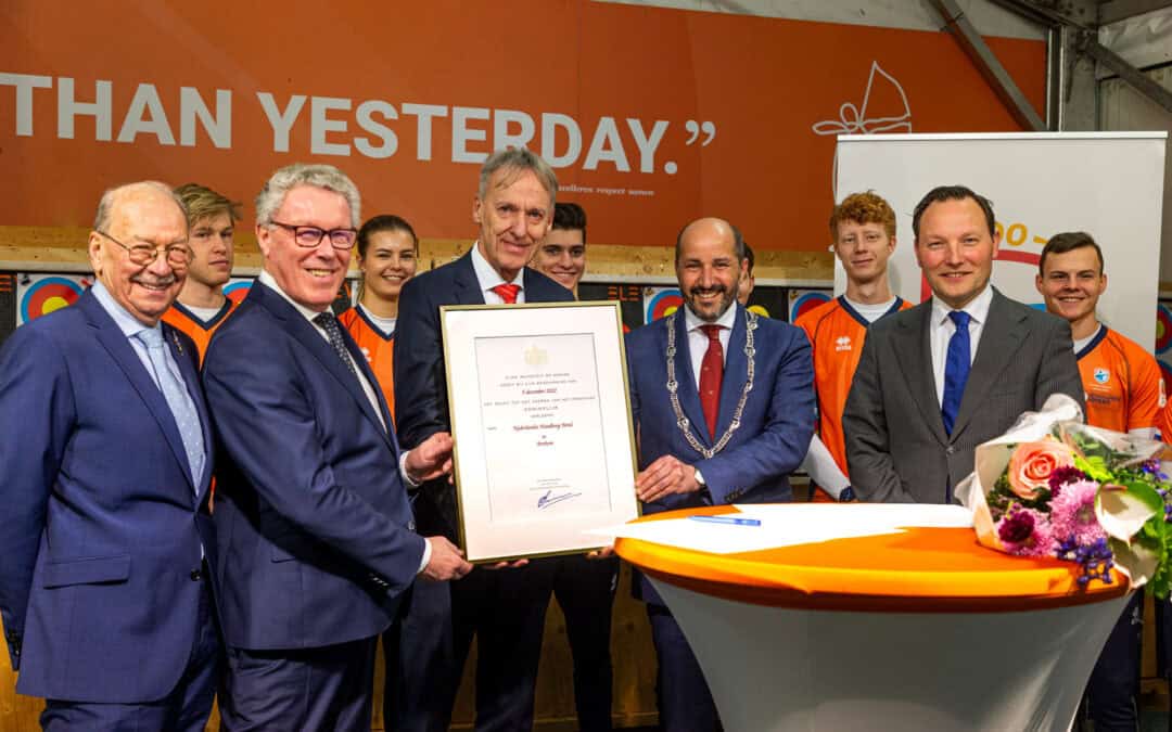 Een groep mensen in formele kledij staat bij elkaar, waarbij één man een ingelijst certificaat vasthoudt. Ze zitten rond een tafel versierd met een boeket bloemen en een boog, wat verwijst naar hun gedeelde passie voor handboogschieten.