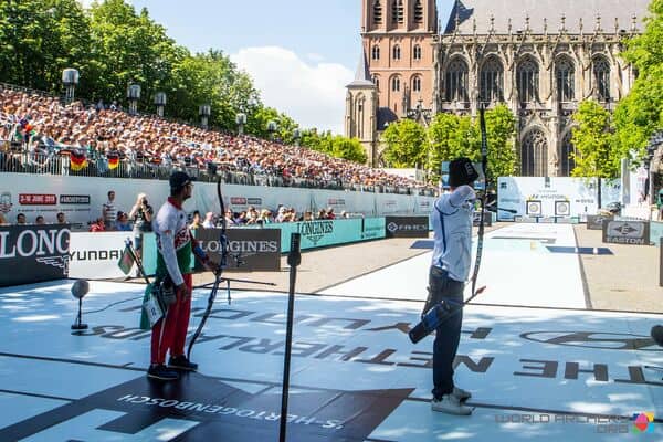 Twee boogschutters in een wedstrijdsetting, waarbij de één zich voorbereidt om een pijl uit zijn boog te schieten en de ander toekijkt. Een grote menigte kijkt toe vanaf tribunes bij een historisch gebouw op de achtergrond, wat bijdraagt aan de sfeer van dit handboogsportevenement.
