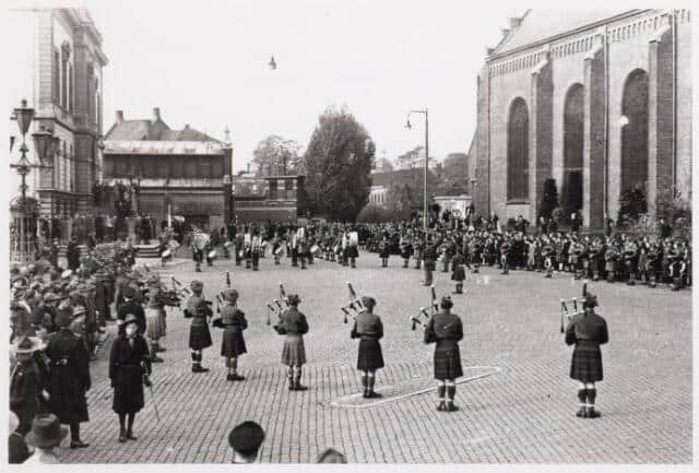 Een groep doedelzakspelers in kilts treedt op in een geplaveide straat vol toeschouwers in de buurt van historische gebouwen, waar een boogschiettentoonstelling de toeschouwers boeit met traditionele handboogschietdemonstraties.