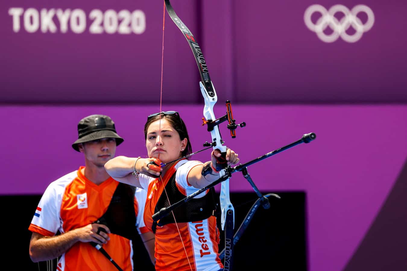 Twee boogschutters in oranje en witte uniformen nemen deel aan de Olympische Spelen van Tokyo 2020. De boogschutter op de voorgrond spant haar boog en richt op een onzichtbaar doelwit. Op de achtergrond zijn de Olympische ringen zichtbaar. Dit moment weerspiegelt de precisie en toewijding van handboogschieten op wereldniveau.