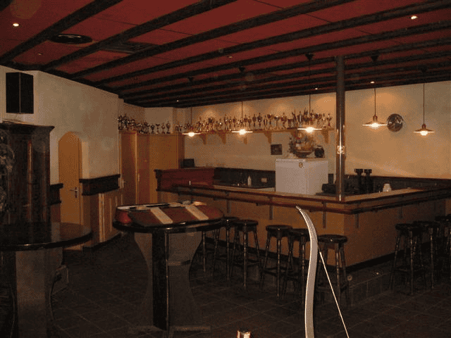 Een slecht verlichte bar is voorzien van houten krukken, een toonbank, plafondverlichting en een verzameling trofeeën op een plank achter de bar, waarvan één prijs opvalt tijdens een boogschietwedstrijd: een bewijs van de liefde van de eigenaar voor boogschieten.