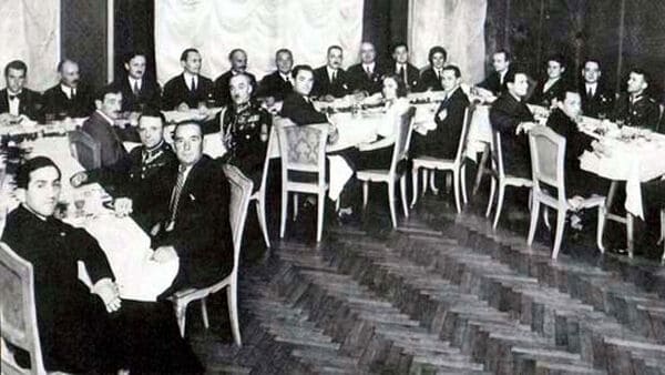Een zwart-witfoto toont een grote groep mannen in formele kledij, zittend aan een lange, U-vormige eettafel tijdens een formeel handboogschieten.
