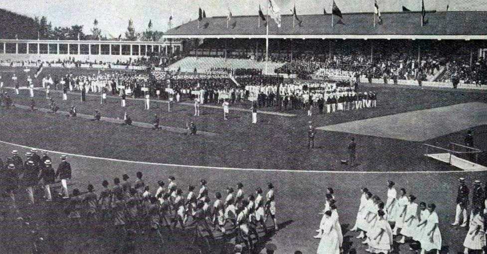 Zwart-witfoto van een grote bijeenkomst van atleten en officials op een stadionveld, mogelijk tijdens een openingsceremonie. Toeschouwers vullen de tribunes en erboven worden nationale vlaggen getoond. Onder de deelnemers bevinden zich deelnemers uit de handboogsport, die hun precisie en vaardigheid met de boog demonstreren.