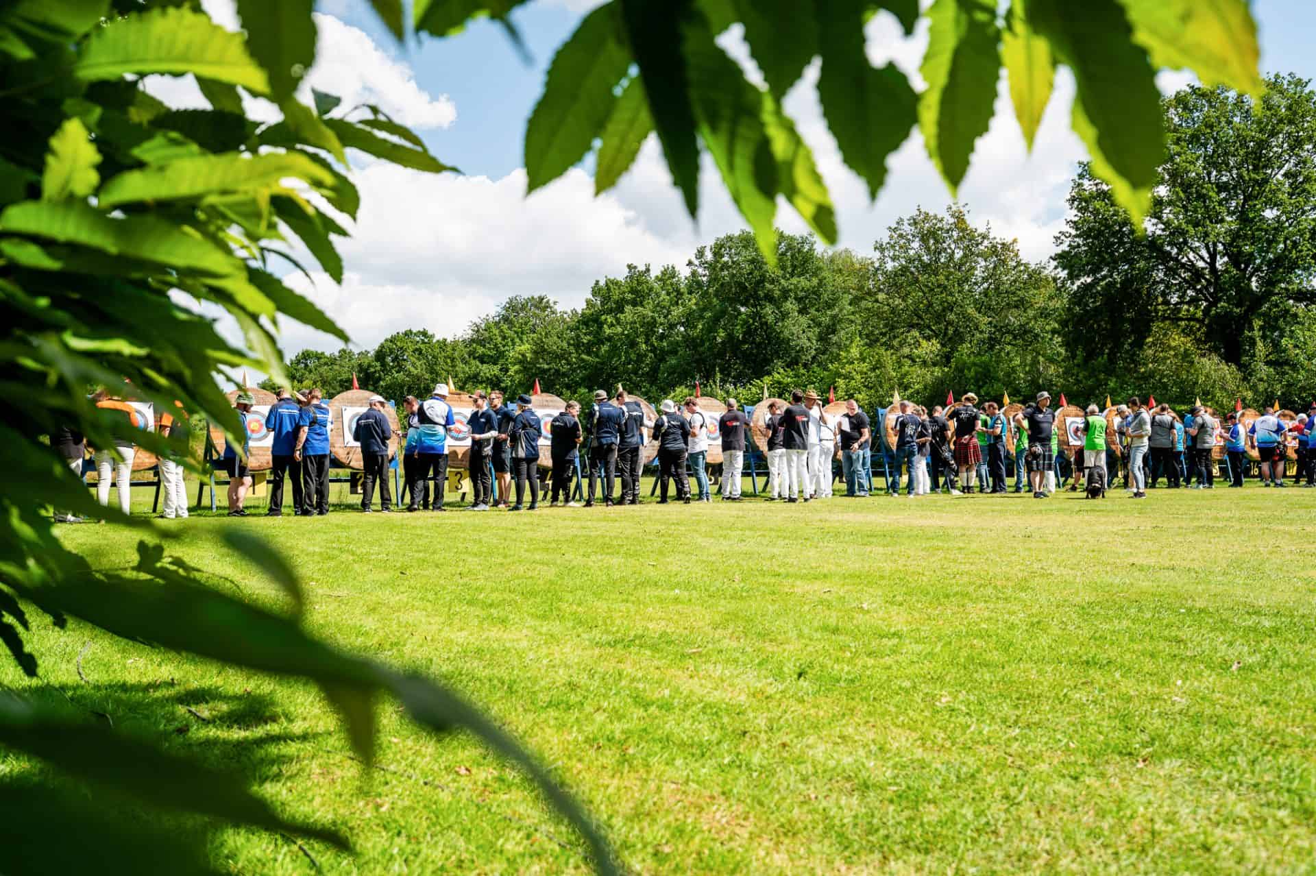 Een grote groep mensen staat in een open grasveld omgeven door bomen, met wat groene bladeren op de voorgrond. Ze lijken samen te komen voor een buitenactiviteit, mogelijk handboogschieten of een ander evenement waarbij handboogsport betrokken is.
