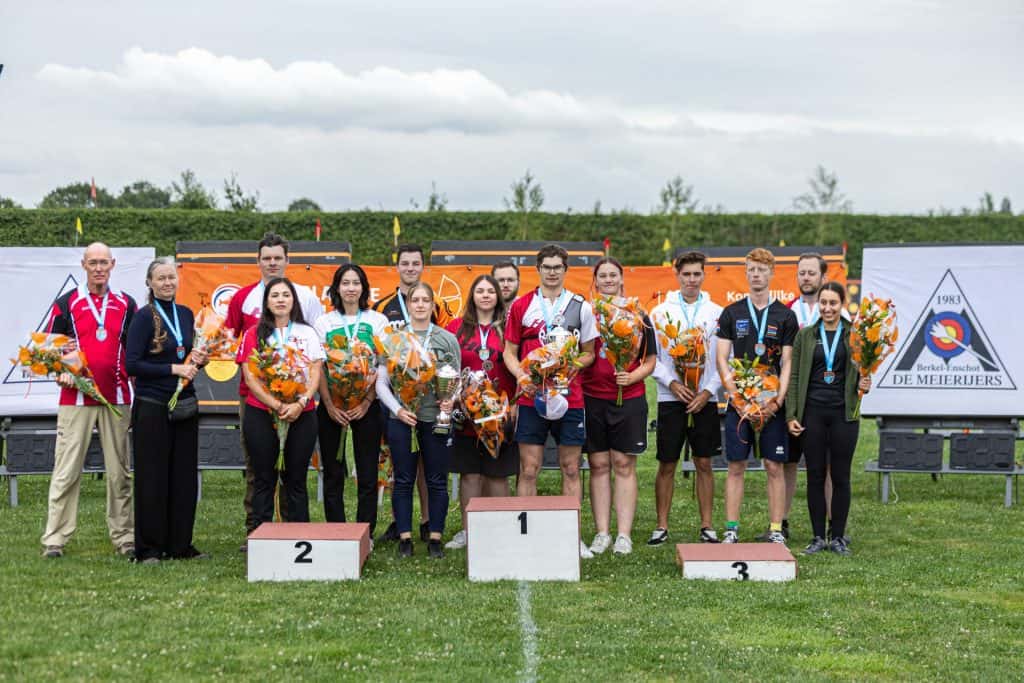 Op en rond podia staat een groep mensen met bloemen en medailles in de hand, waarmee ze aangeven dat ze winnaars zijn van een handboogsportwedstrijd. De achtergrond toont spandoeken en een bewolkte hemel.